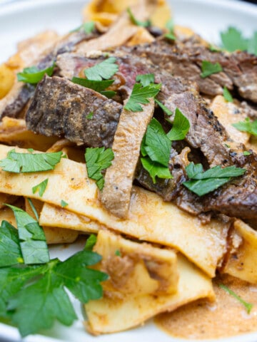 Beef stroganoff with flank steak, mushrooms and pasta on a white plate garnished with parsley.