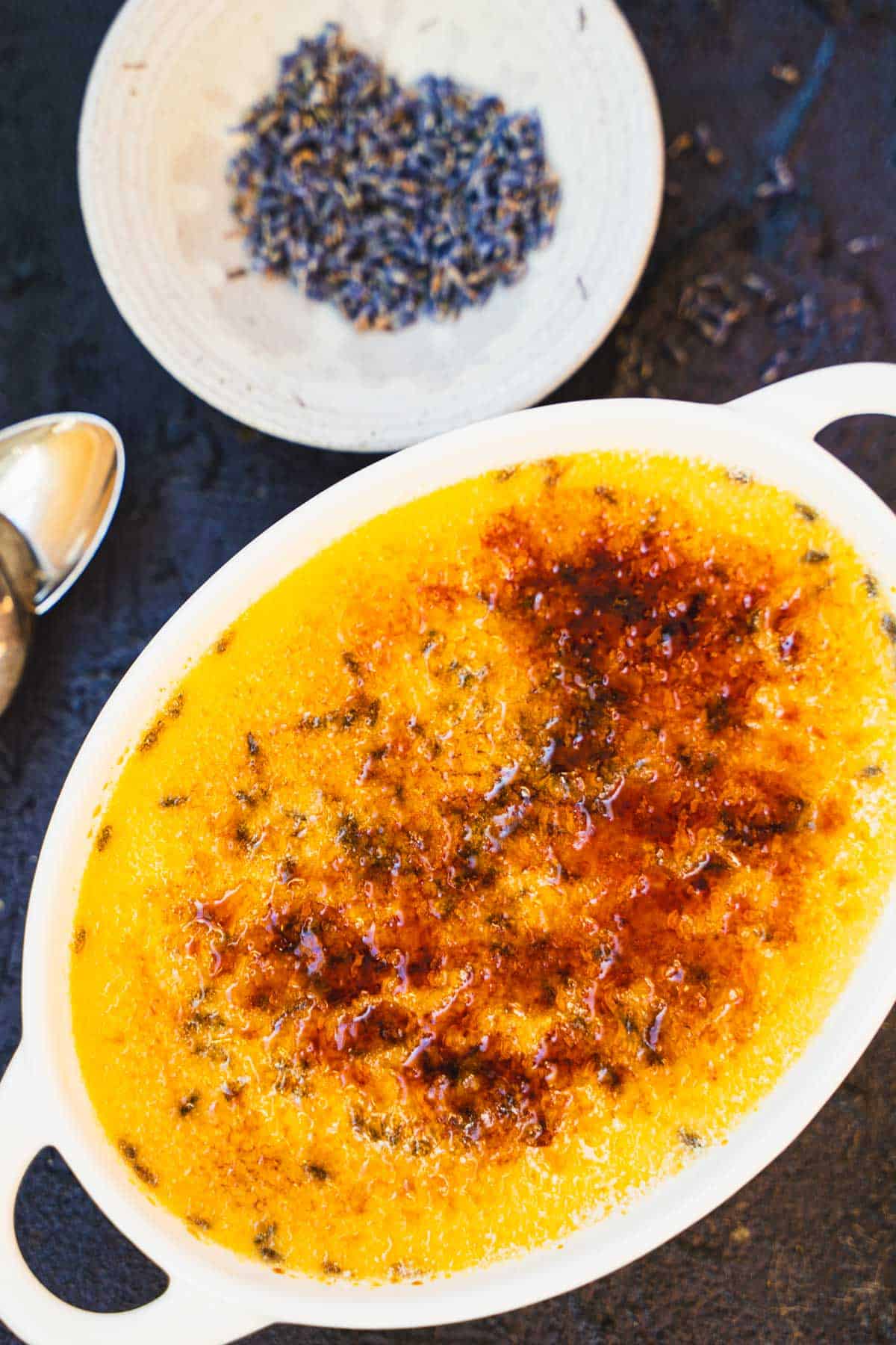 Easy lavender Creme Brulee for 2 in a white ramekin next to two spoons and a bowl of lavender