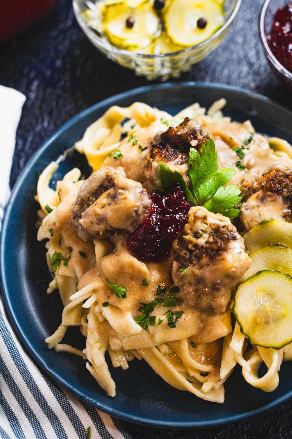 swedish meatballs and a sauce made with sourcream over a bed of buttered noodles on a blue plate garnished with lingonberry jam and pickled cucumbers.