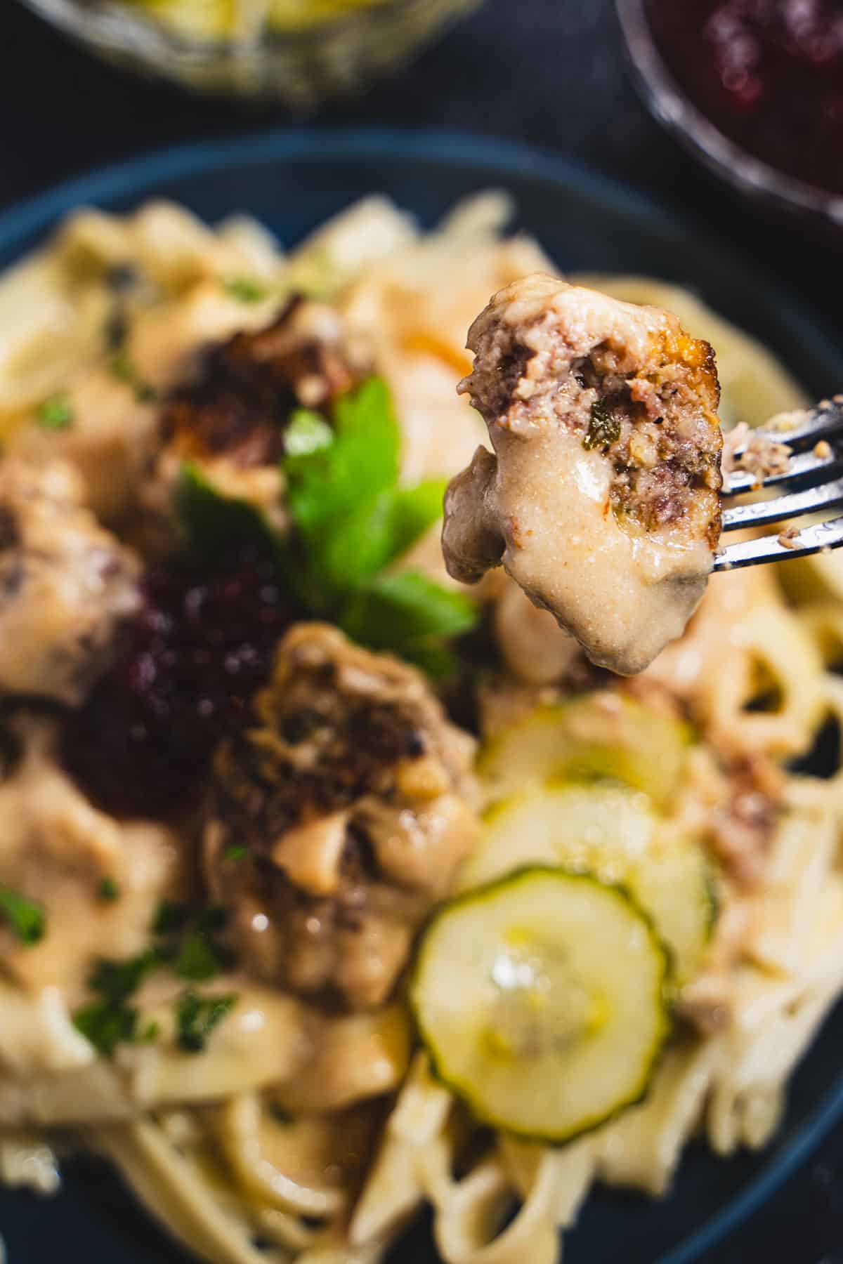 swedish meatball on a fork over a plate of swedish meatballs and a sauce made with sourcream over a bed of buttered noodles on a blue plate garnished with lingonberry jam and pickled cucumbers.