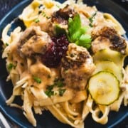 Swedish meatballs and sauce on a blue plate garnished with lingonberry sauce and pickled cucumbers.