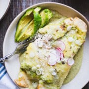 one pan chili verde enchiladas with chicken and green sauce in a white bowl on a wood table next to bowls of spanish rice and cotija cheese.