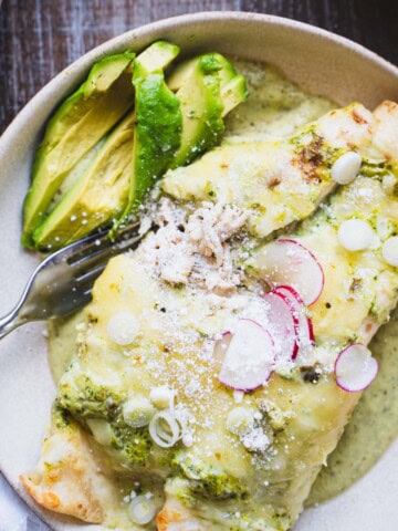 one pan chili verde enchiladas with chicken and green sauce in a white bowl on a wood table next to bowls of spanish rice and cotija cheese.