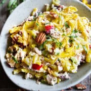 Cold tuna pasta salad with apples and pecans tailored for two in a green bowl on a wood table surrounded by dry pasta, lemon, onion and a small marble bowl of salt and a tarragon sprig.