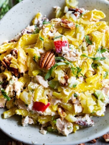 Cold tuna pasta salad with apples and pecans tailored for two in a green bowl on a wood table surrounded by dry pasta, lemon, onion and a small marble bowl of salt and a tarragon sprig.