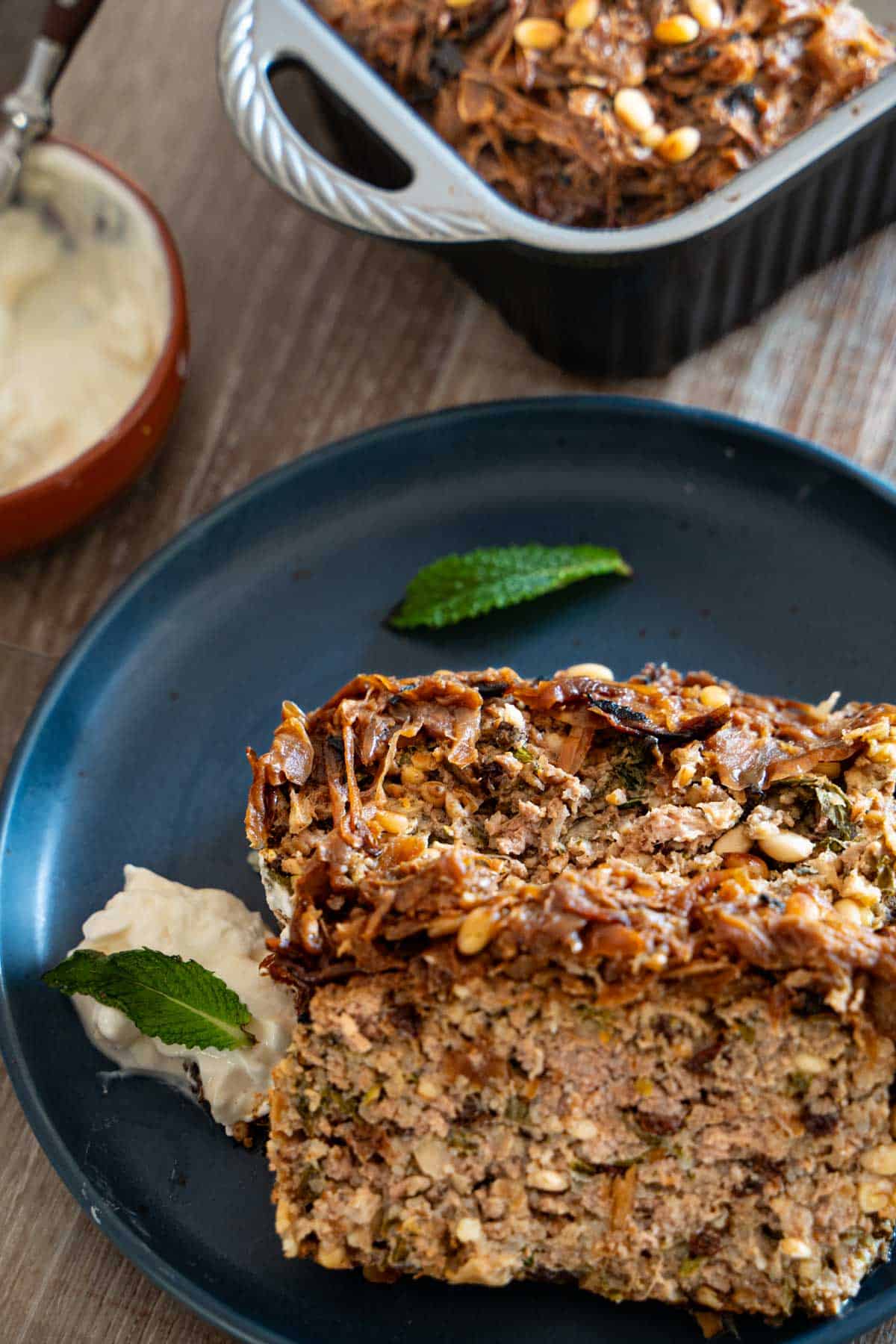 Two slices of Lamb meatloaf on a blue plate the rest in the loaf pan served alongside hot honey yogurt.