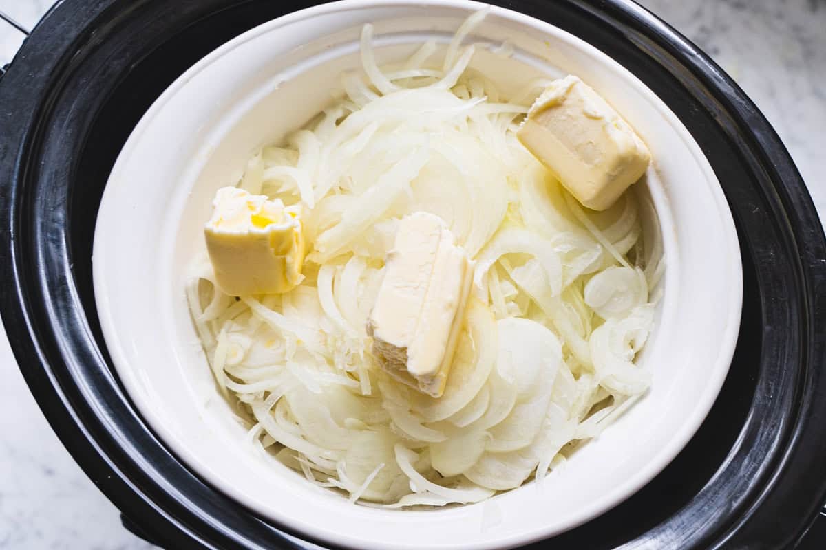 small white oven safe pyrex dish inside a large slow cooker bowl with white onions and pads of butter to make caramelized onion topping for lamb meatloaf.