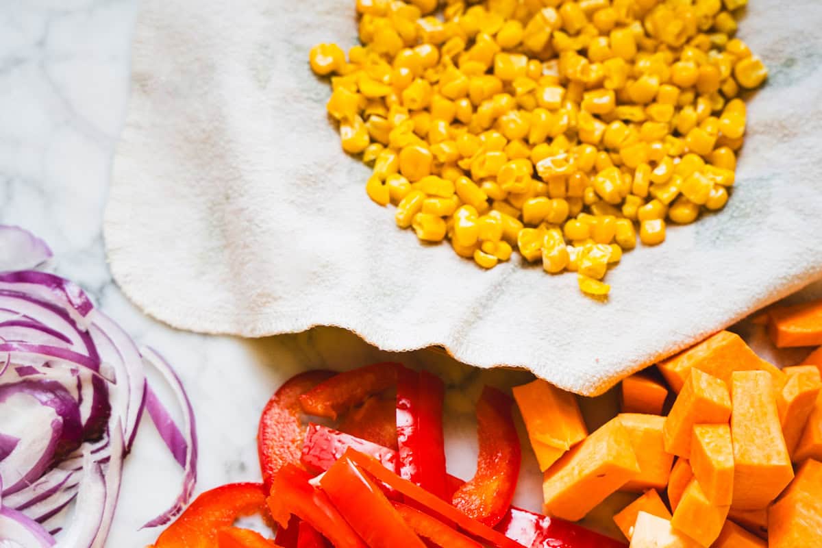 Vegetable bowl vegetables cut and placed on white counter top alongside frozen corn on a paper towel.