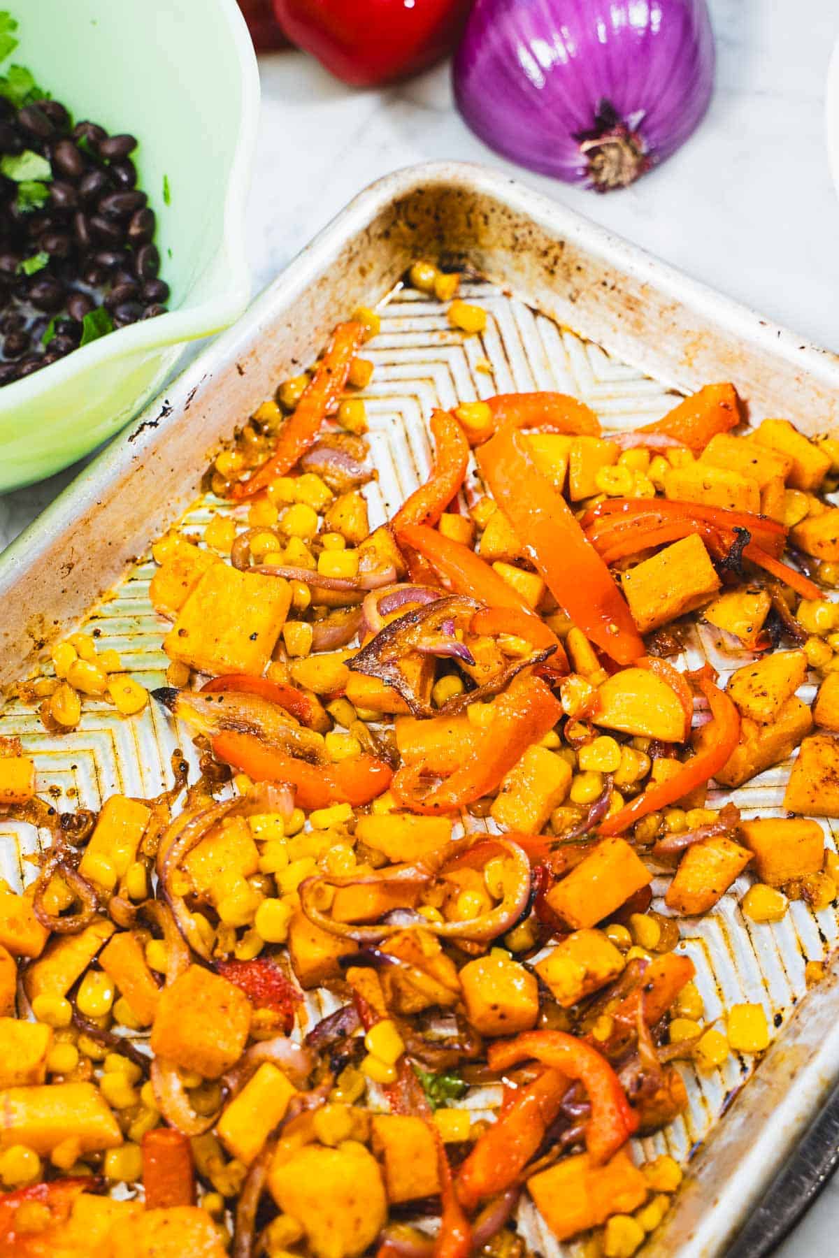 Roasted vegetables for roasted vegetable fajita bowl with hot honey sauce on baking sheet next to seasoned black beans in a green bowl.