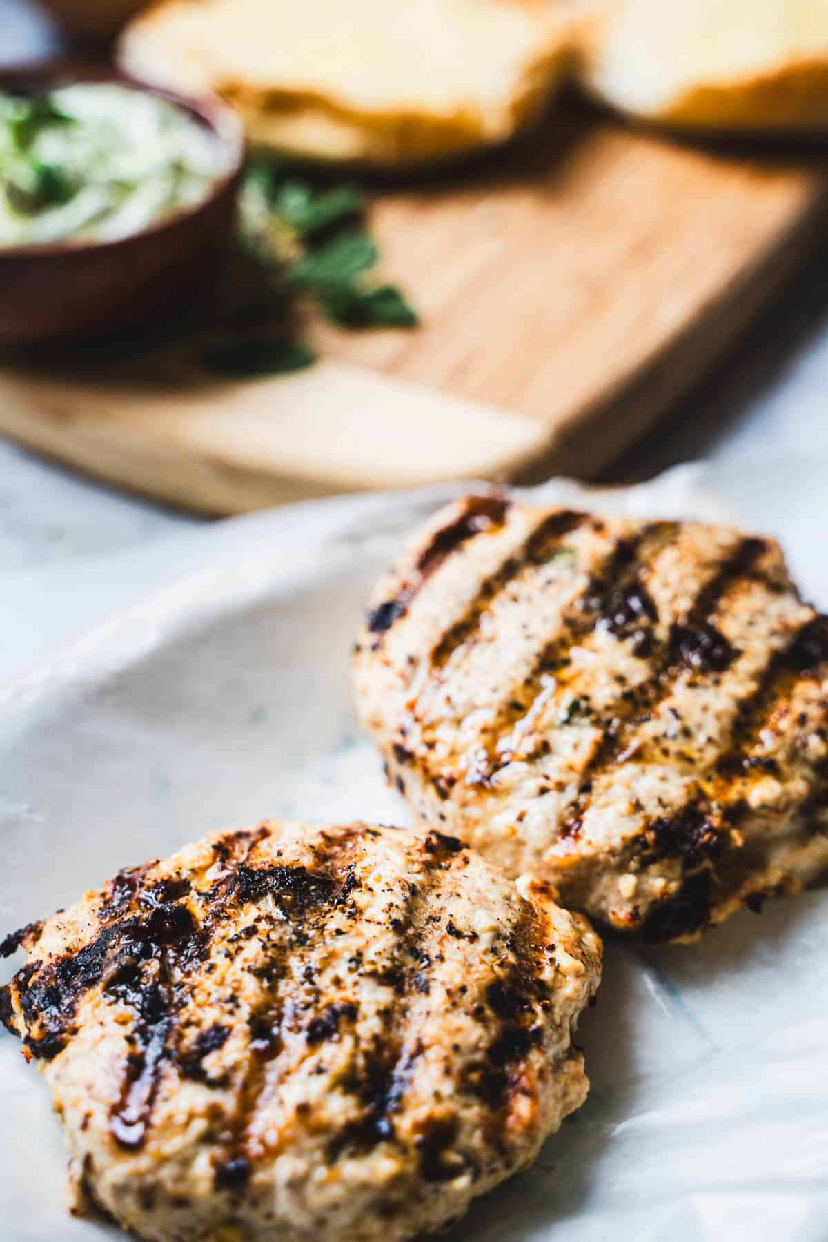 grilled chicken patties resting on parchment paper next to a wooden cutting board with toasted buns.