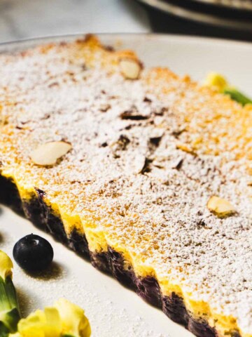 Blueberry almond lemon cake on a white plater dusted with powdered sugar and sprinkled with almonds.