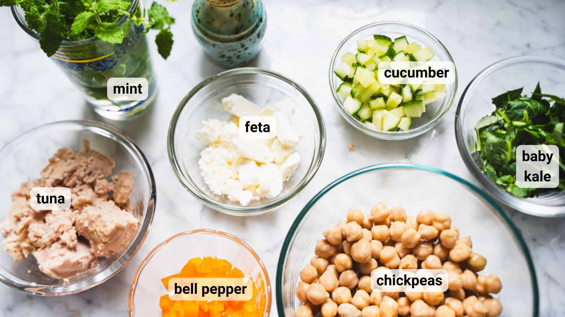 Kale chickpea cucumber feta salad with tuna stuffed avocado ingredients in glass bowls on white marble.