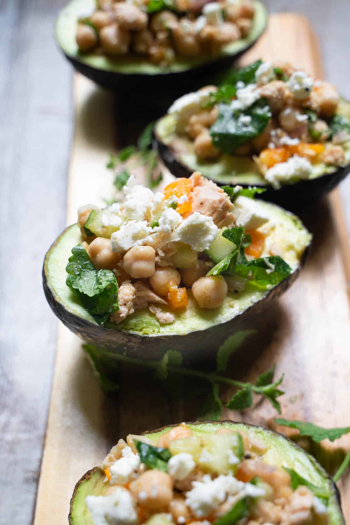 Four Stuffed avocados with kale chickpea salad with tuna filling on a wooden board in a straight line garnished with mint, feta and cucumber.