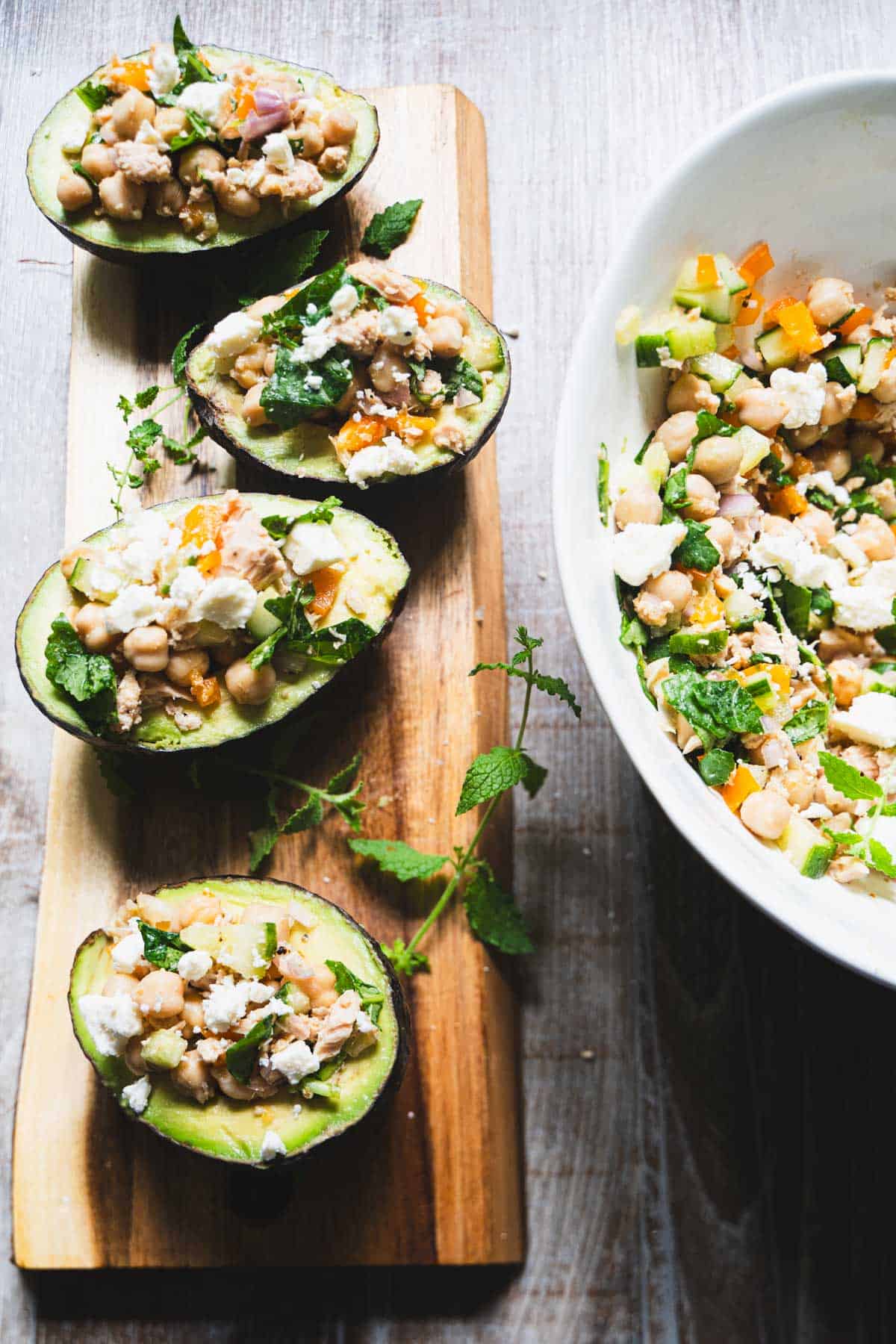 Stuffed Avocados with kale chickpea salad with tuna on a blond wood board next to a white salad bowl.