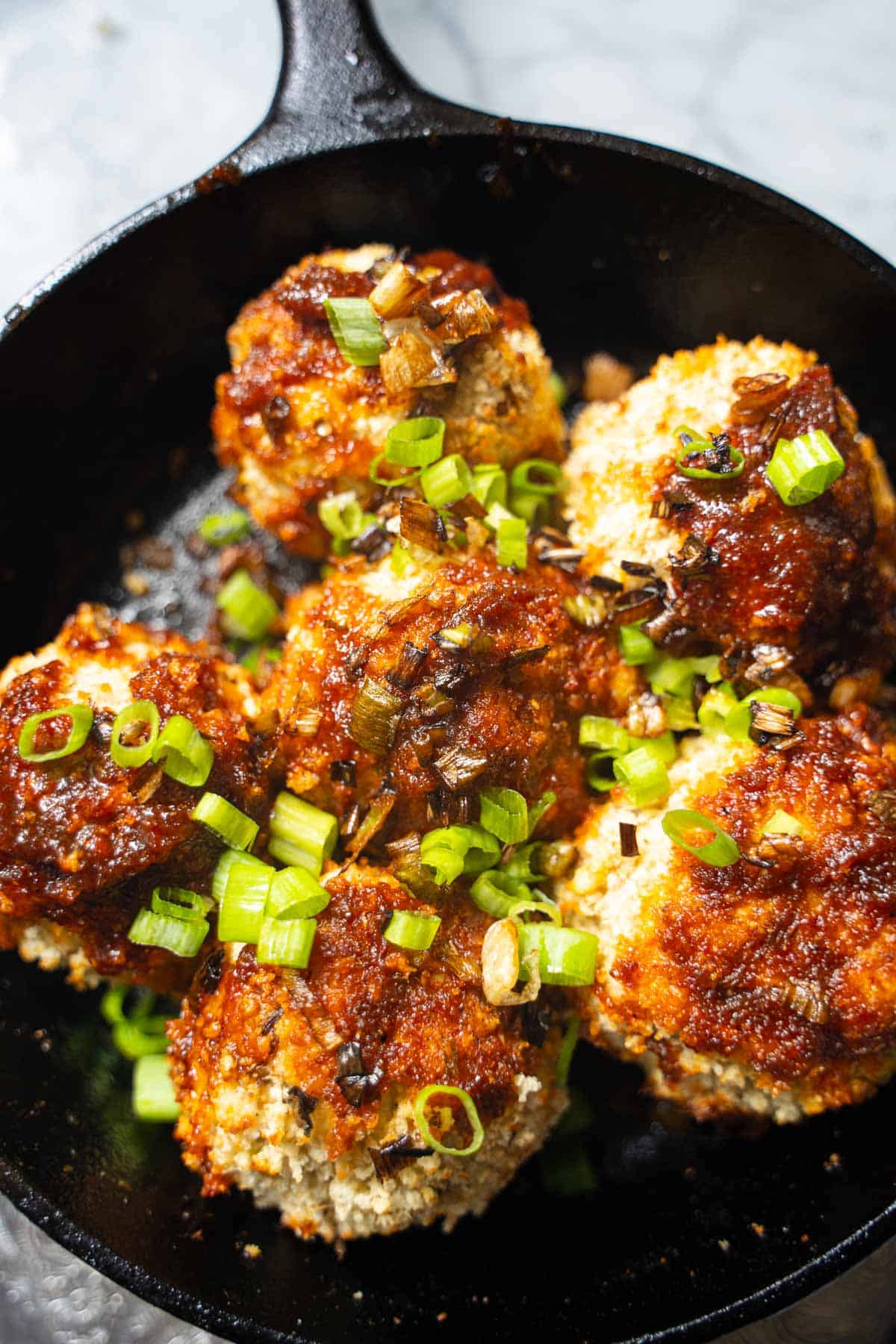 Asian eggplant meatballs with gochujang glaze in a black cast iron skillet garnished with raw and fried scallions.