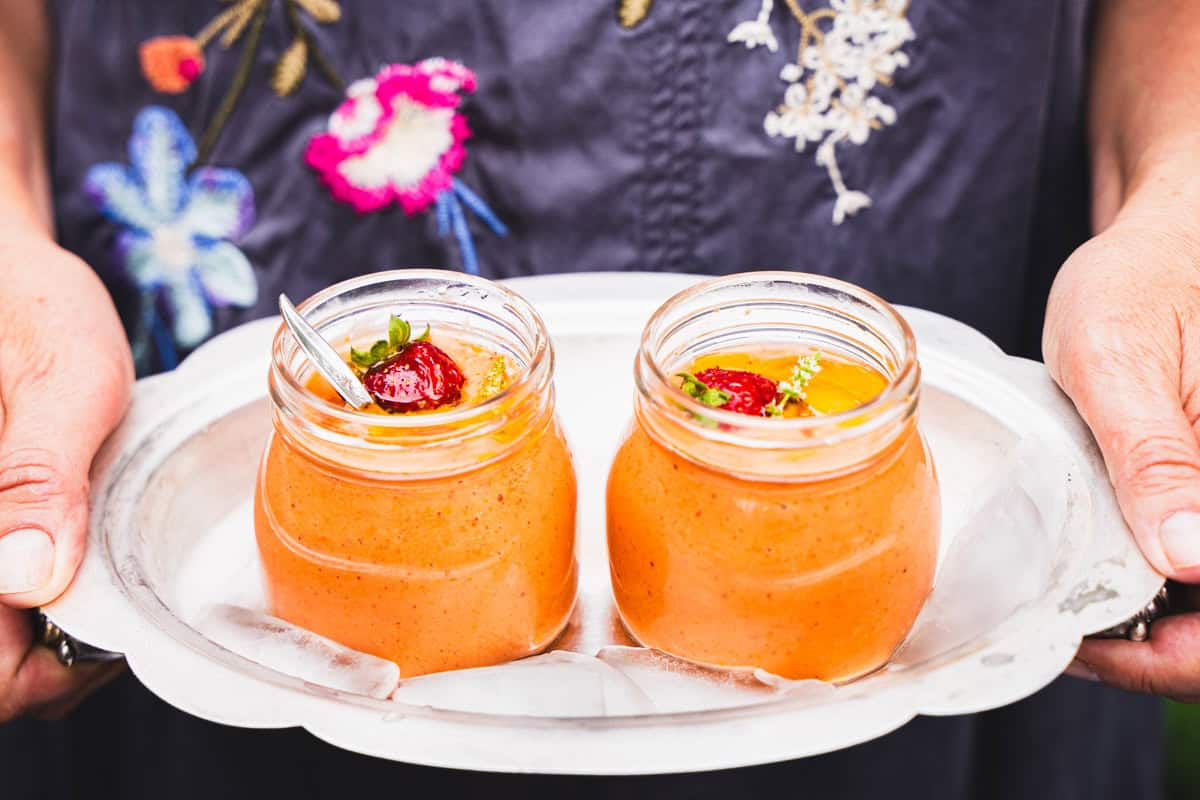 Easy Strawberry and Heirloom Gazpacho in individual sized mason jars on a silver plater held by a women in a flower dress.