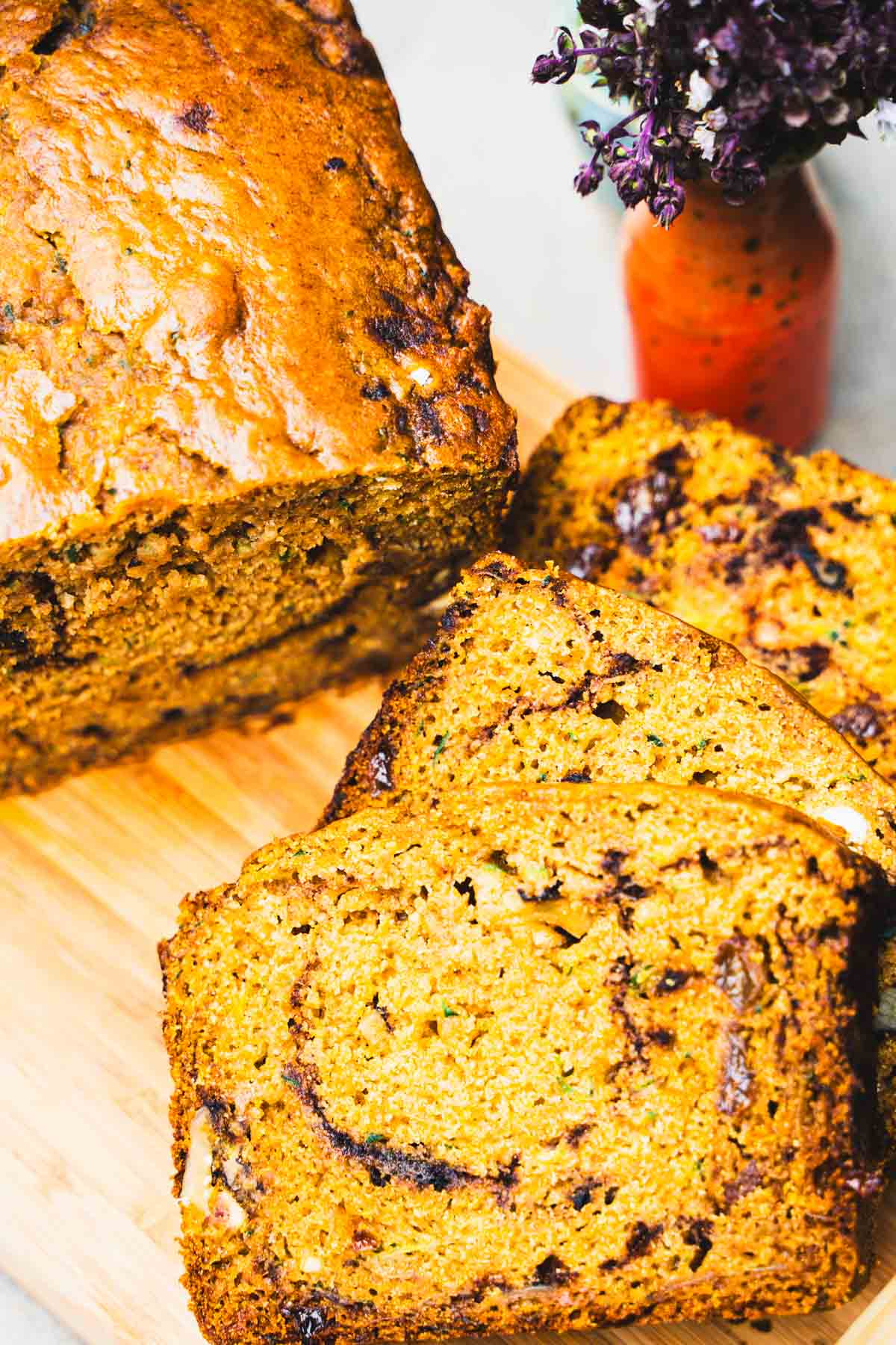 Gluten free zucchini bread sliced on a wooden cutting board next to a small vase of purple flowers.