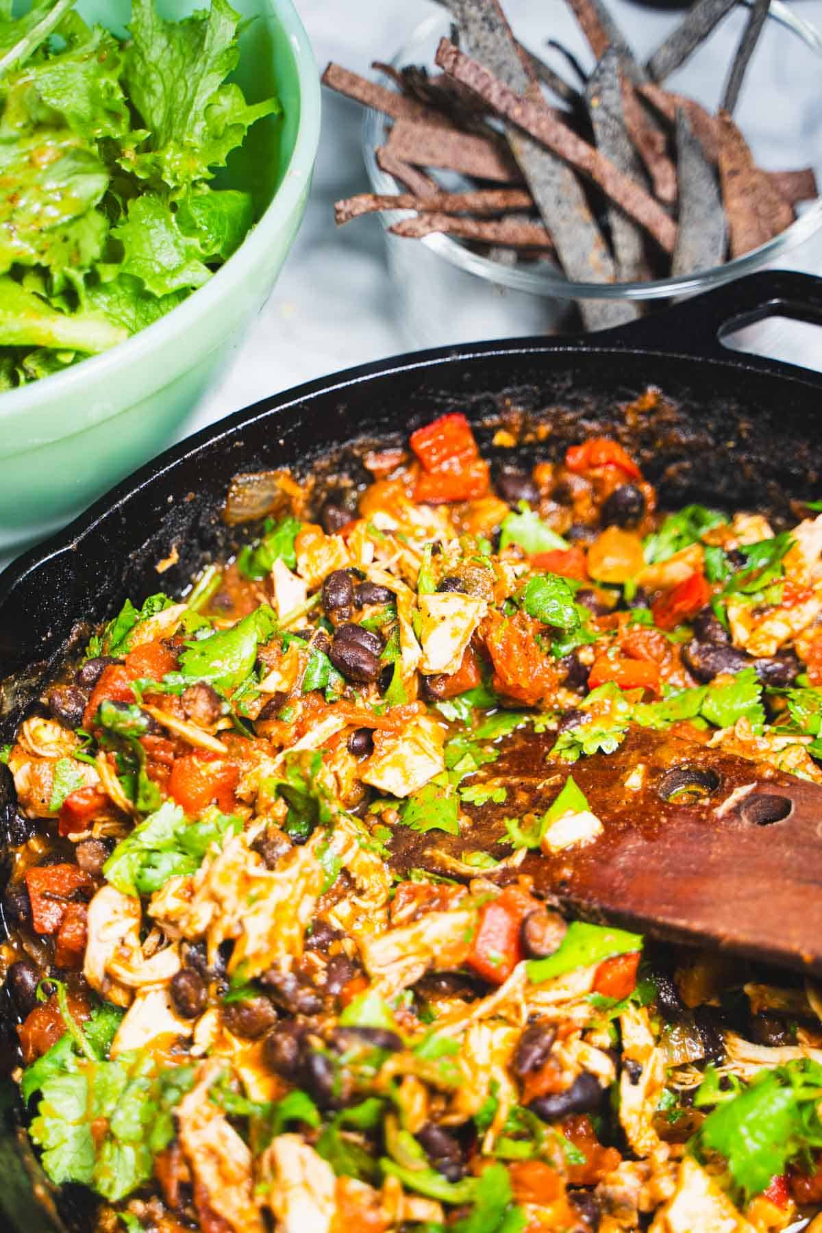 Chicken taco salad chicken mixture in a cast iron skillet with cilantro mixed in, a green bowl of mixed greens with gluten free salad dressing and baked gluten free corn tortilla strips in a clear glass bowl on a white marble counter top.