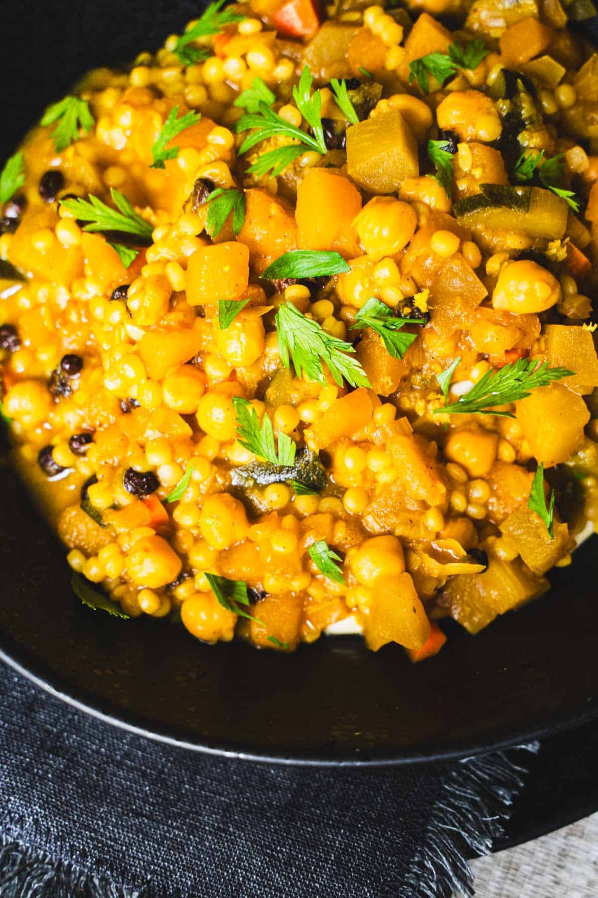 Vegetarian butternut squash, chickpea, zucchini and eggplant tagine with couscous in a black bowl placed on a black denim napkin.