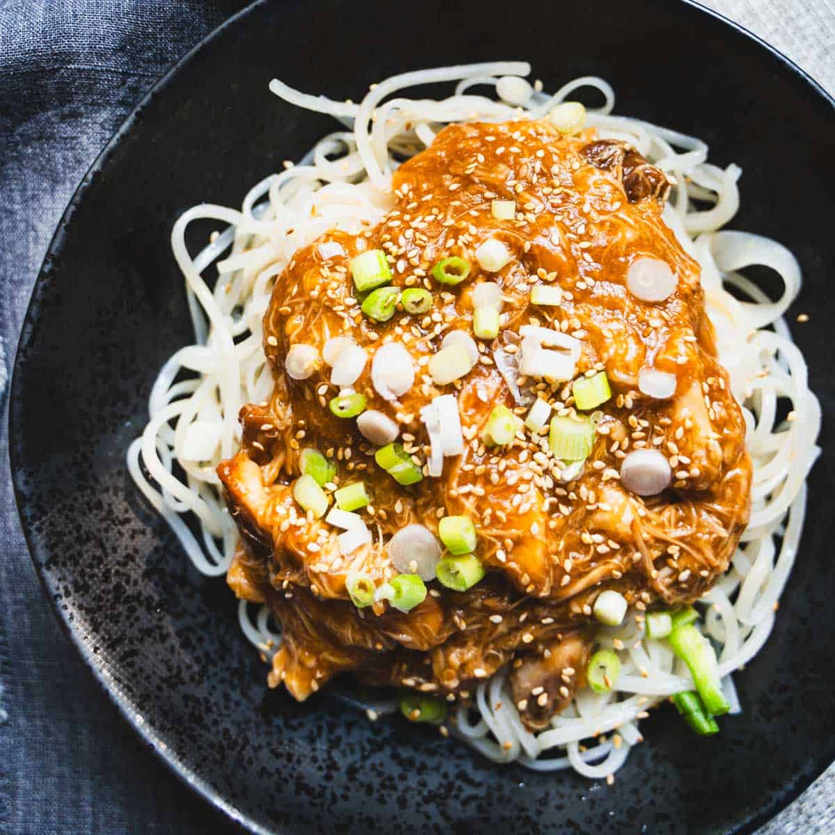Crock pot chicken and noodles with huli huli sauce in a black bowl garnished with sesame seeds and thinly sliced green onions.
