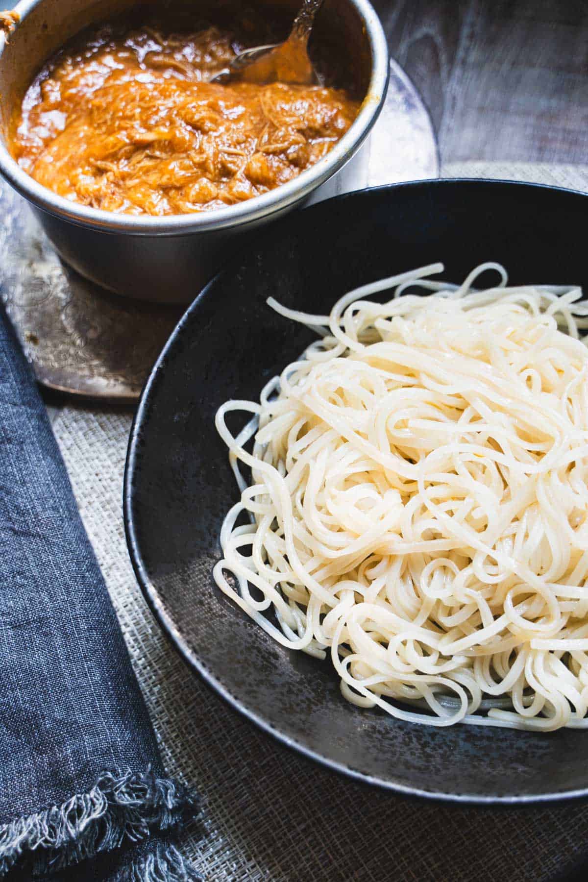 Cooked rice noodles in a black bowl alongside the huli huli chicken on a small baking dish.