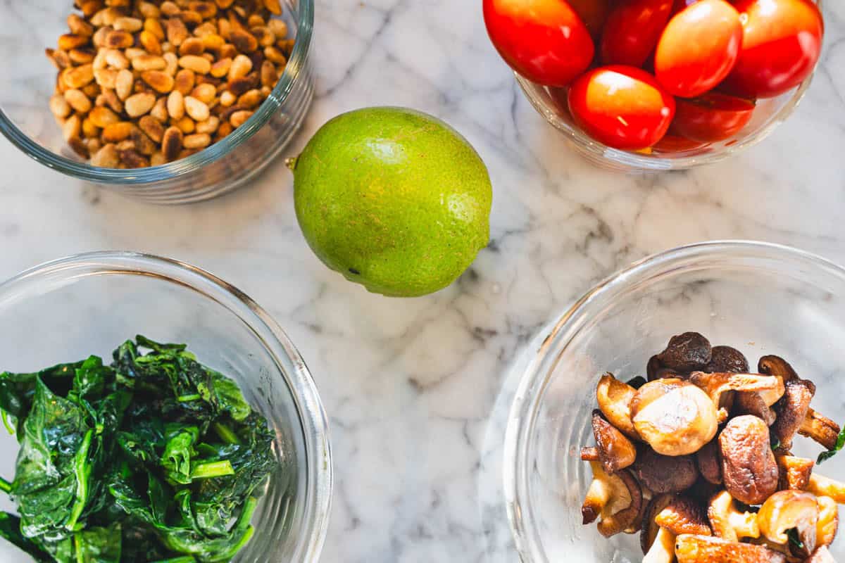 Mushrooms, pine nuts, cherry tomatoes, spinach and lime on a white counter to garnish the red thai coconut curry soup recipe.