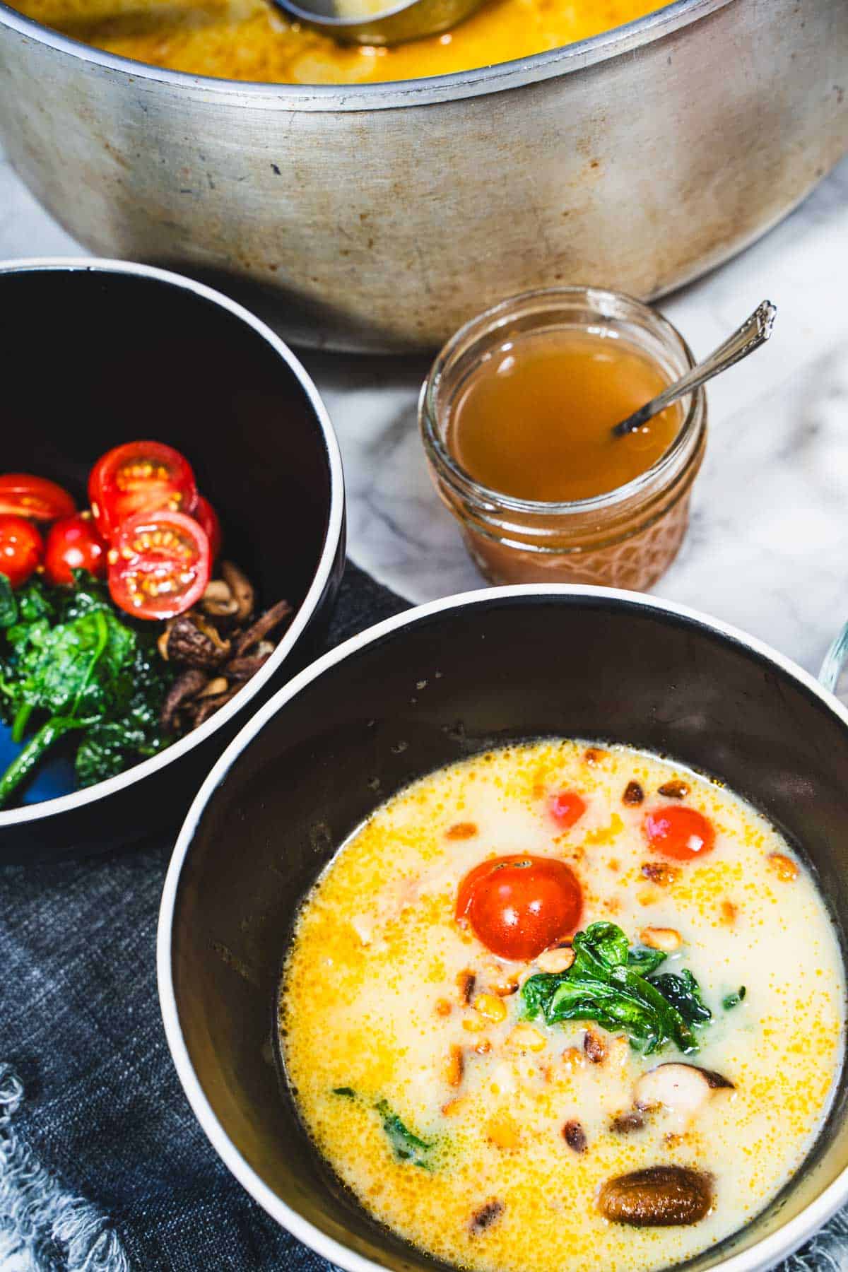 Red Thai Coconut Spinach Tomato Curry broth ladled over garnishes in one bowl, pot of coconut curry broth, jar of tomato essence on white marble counter.