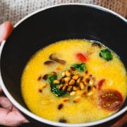 Red Thai Coconut, Spinach, Tomato Curry Soup in a black bowl with a white rim held in a women's hands wearing a beige sweater.