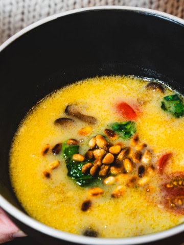 Red Thai Coconut, Spinach, Tomato Curry Soup in a black bowl with a white rim held in a women's hands wearing a beige sweater.