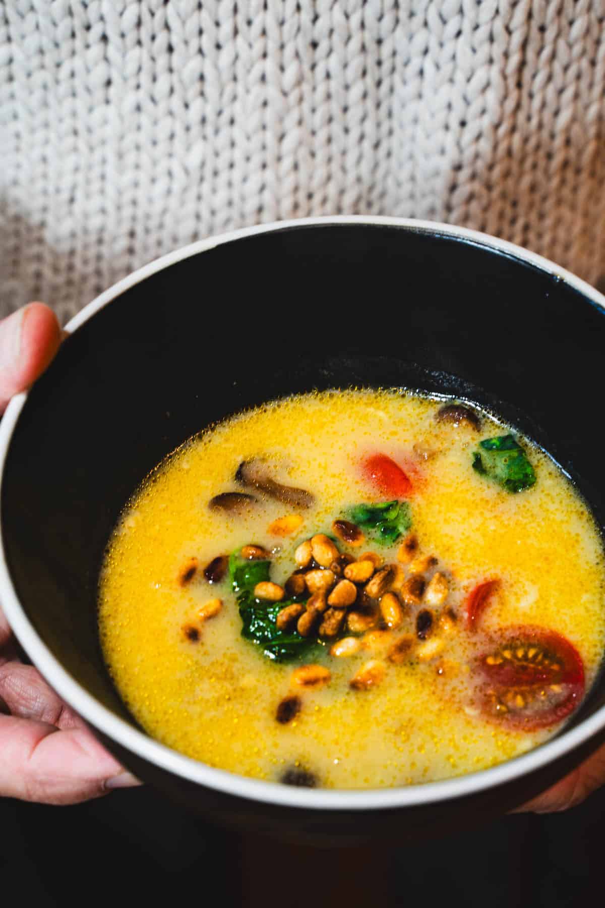 Red Thai Coconut, Spinach, Tomato Curry Soup in a black bowl with a white rim held in a women's hands wearing a beige sweater.