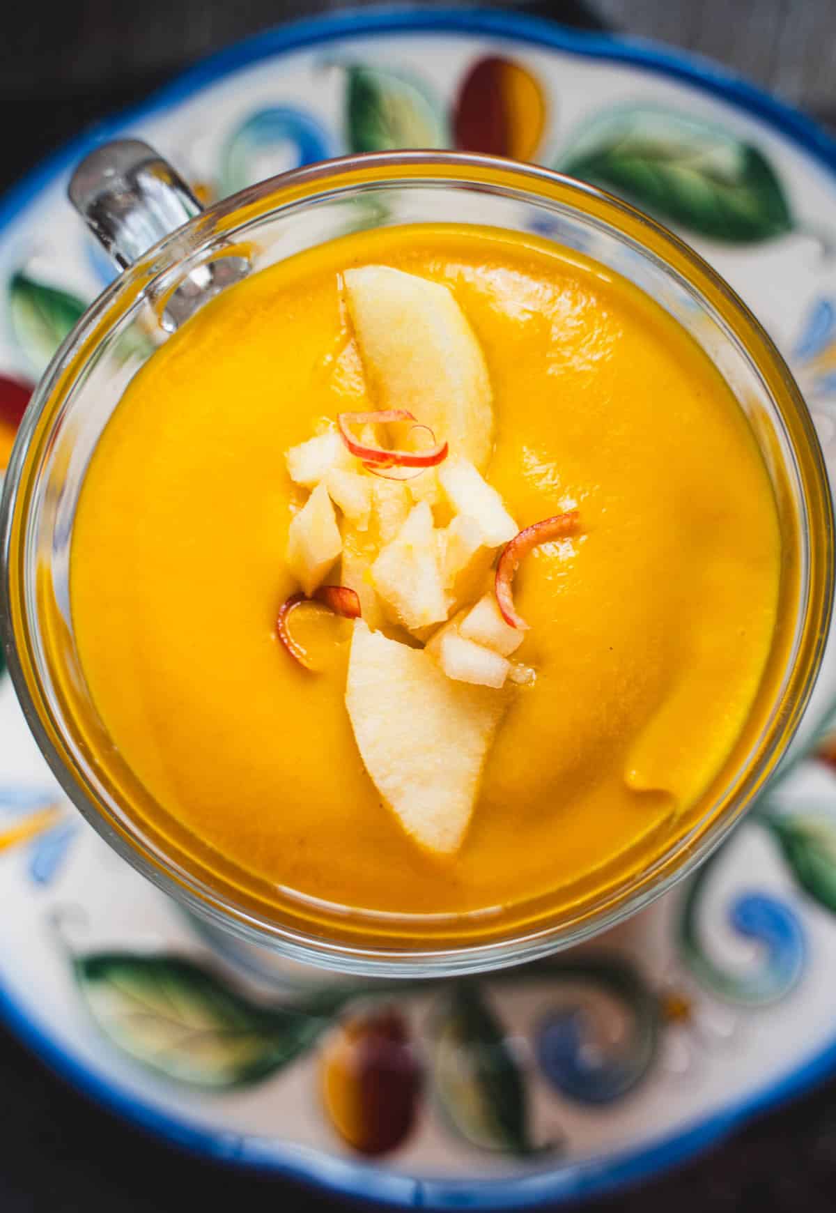 apple autumn squash soup in a glass mug on a flowered plate garnished with diced apple.