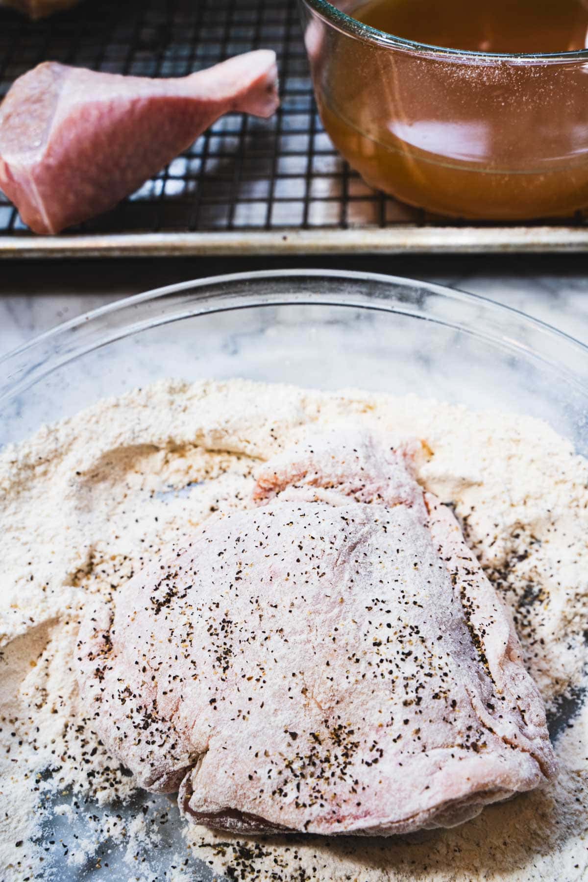 A shallow pie plate with seasoned flour ready for dredging chicken alongside a wire rack inserted into a baking sheet with chicken and apple cider on it.