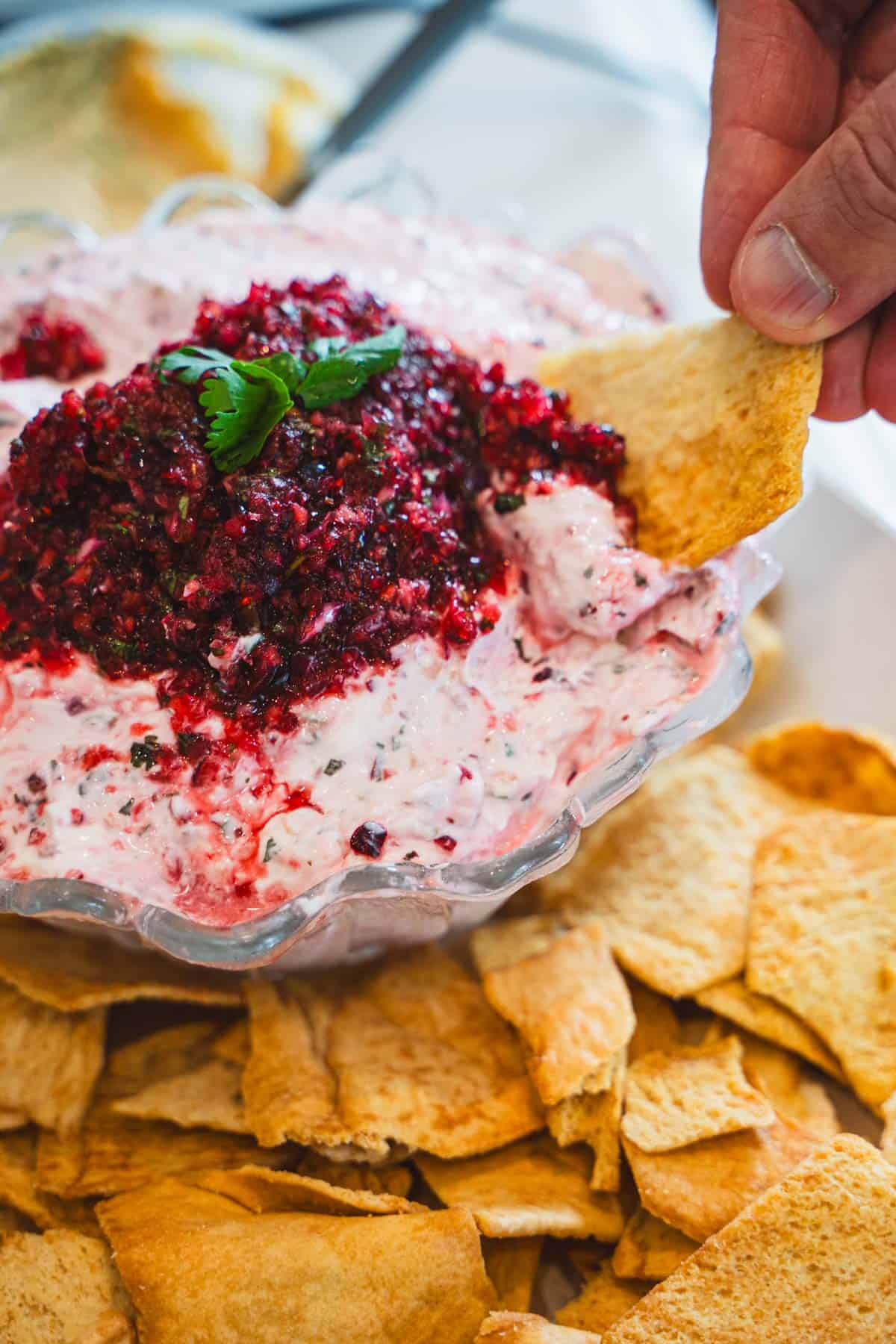 Cranberry and Jalapeno dip with cream cheese in a clear glass serving bowl surrounded by pita chips with a hand dipping a pita chip into the bowl.