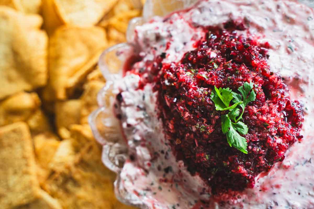 Cranberry and Jalapeno dip with cream cheese in a clear glass serving bowl surrounded by pita chips.