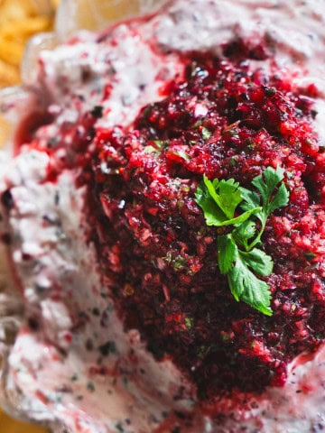Cranberry and Jalapeno dip with cream cheese in a clear glass serving bowl surrounded by pita chips.