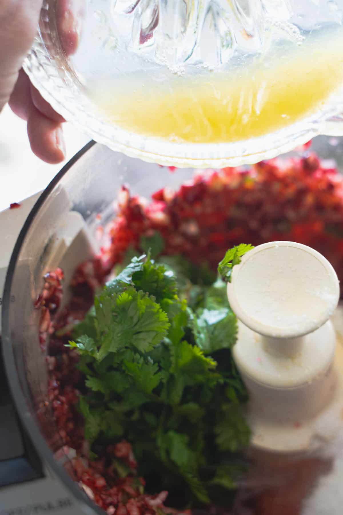 Pouring orange juice into food processor bowl with finely chopped cranberries, cilantro leaves and sugar.