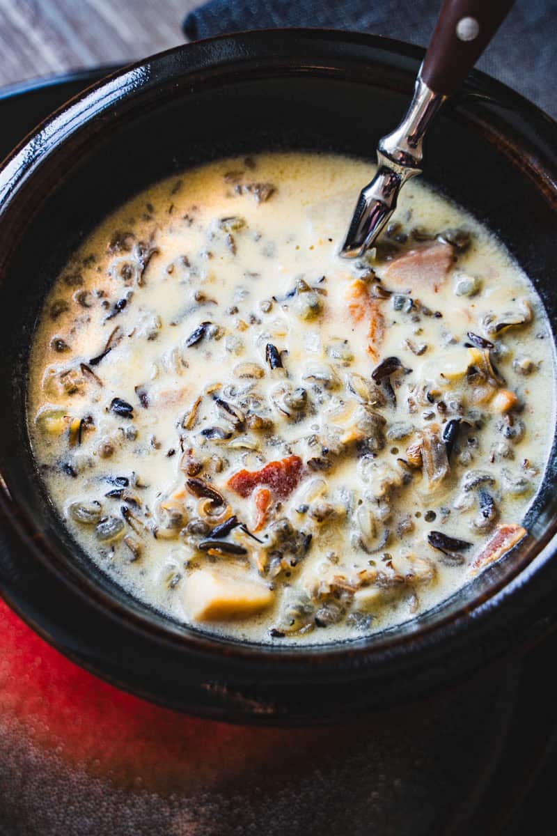 creamy wild rice soup with bacon and ham in a brown bowl sitting on a red and brown plate.
