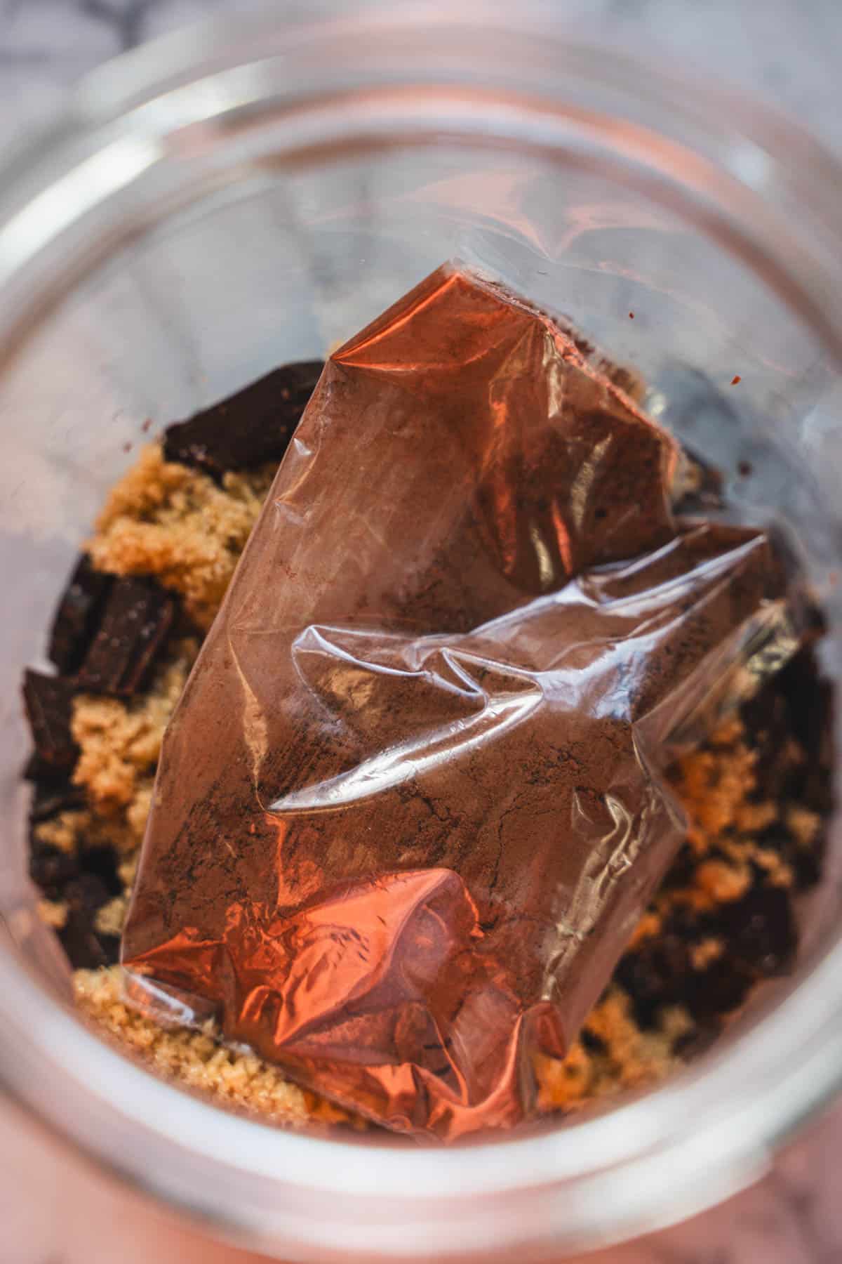 Cocoa powder in a small plastic bag in glass jar on top of dark hot chocolate mix ingredients.