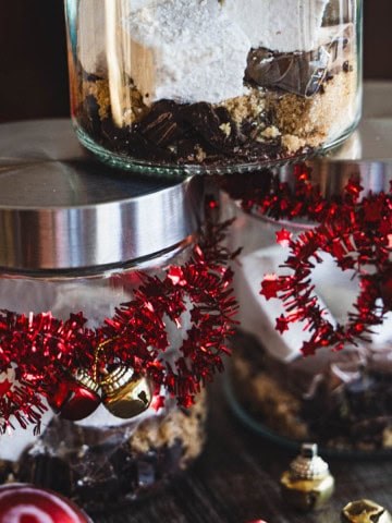 Dark hot chocolate mix and homemade marshmallows in a jar decorated with red garland and small gold and red bells.