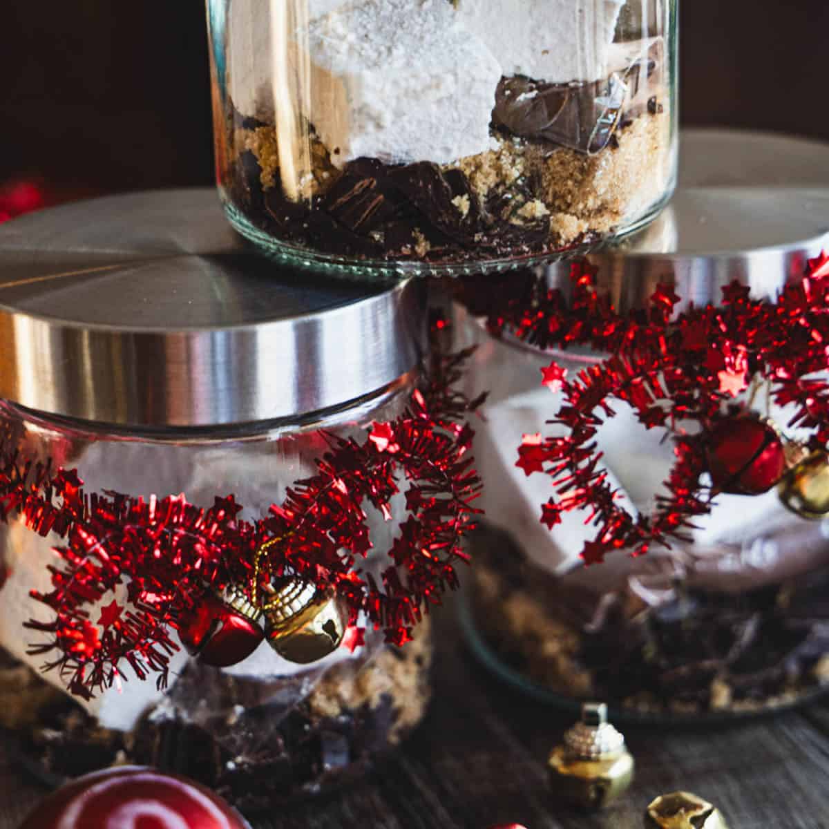 Dark hot chocolate mix and homemade marshmallows in a jar decorated with red garland and small gold and red bells.