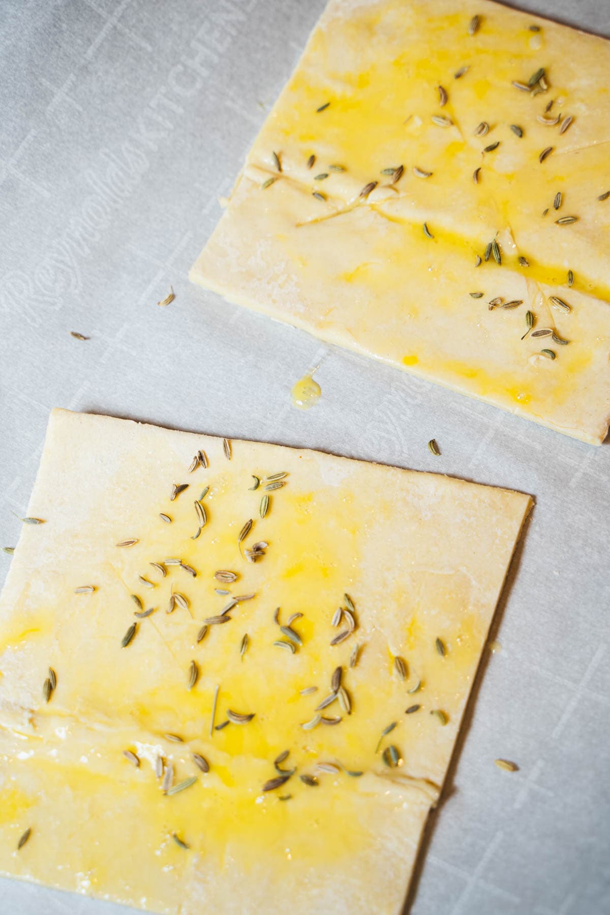 two puff pastry squares on parchment lined baking sheet with egg wash and fennel seeds sprinkled on top.