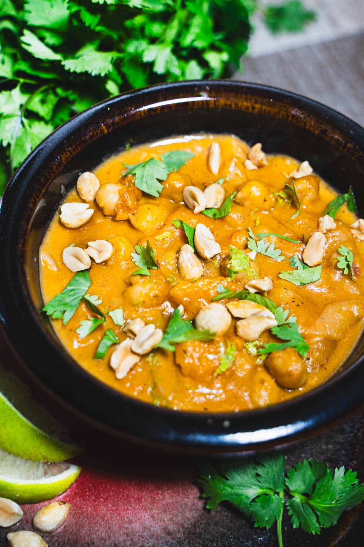 Peanut butter and sweet potato coconut milk soup in a brown bowl surrounded by bunches of cilantro and slices of lime.