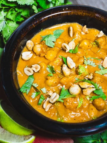 Peanut butter and sweet potato coconut milk soup in a brown bowl surrounded by bunches of cilantro and slices of lime.