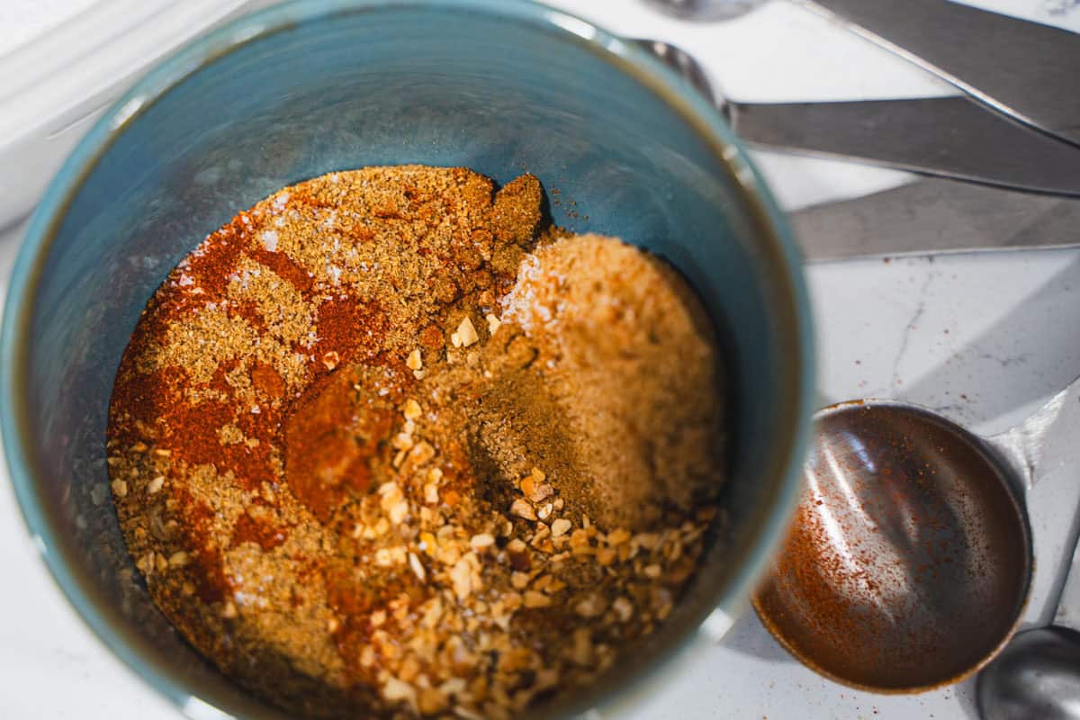 BBQ Ribs dry spice rub in a blue bowl with measuring spoons on white marble counter.