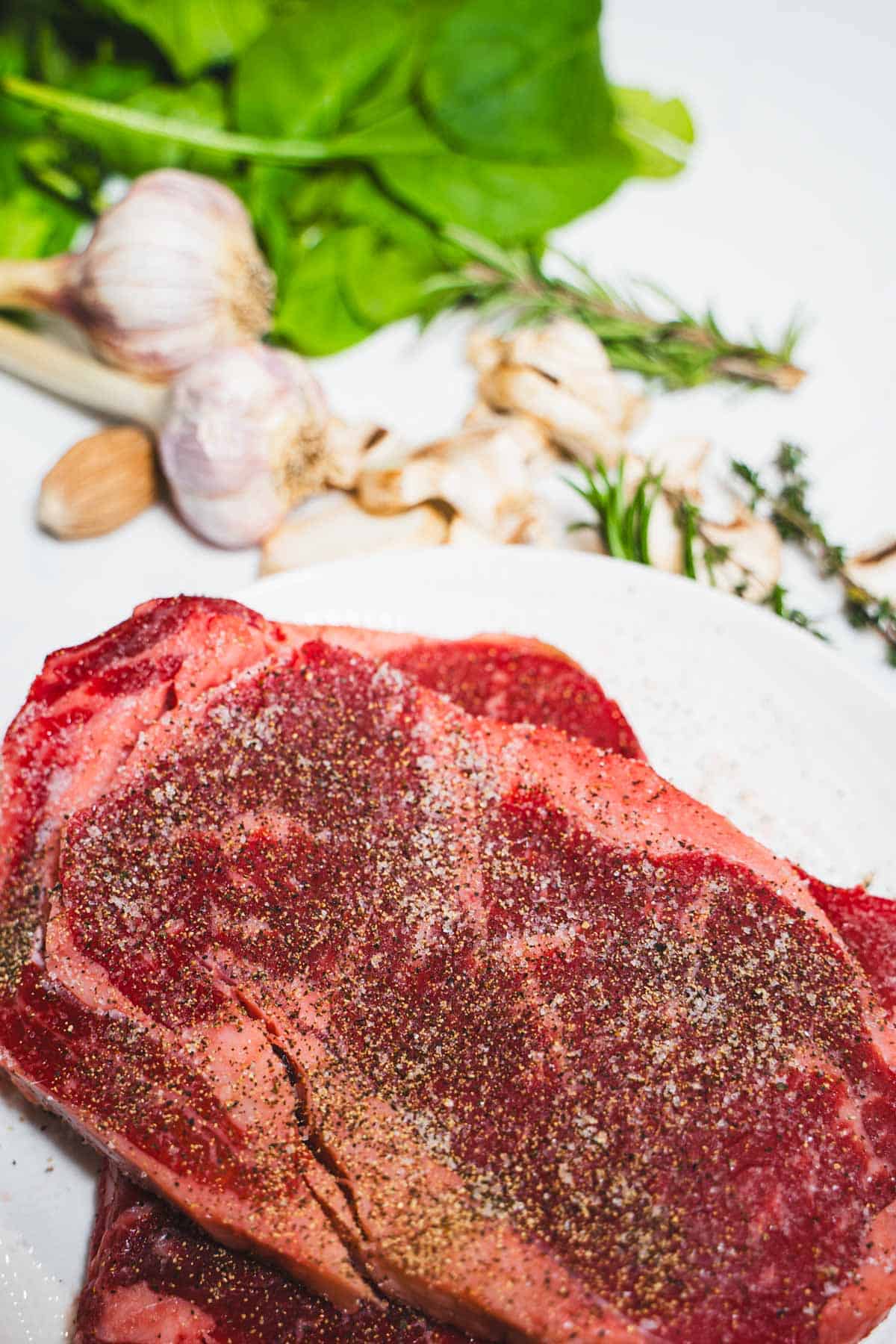 Salt and pepper seasoned ribeye with spinach, mushrooms and herbs and garlic resting on white marble counter.