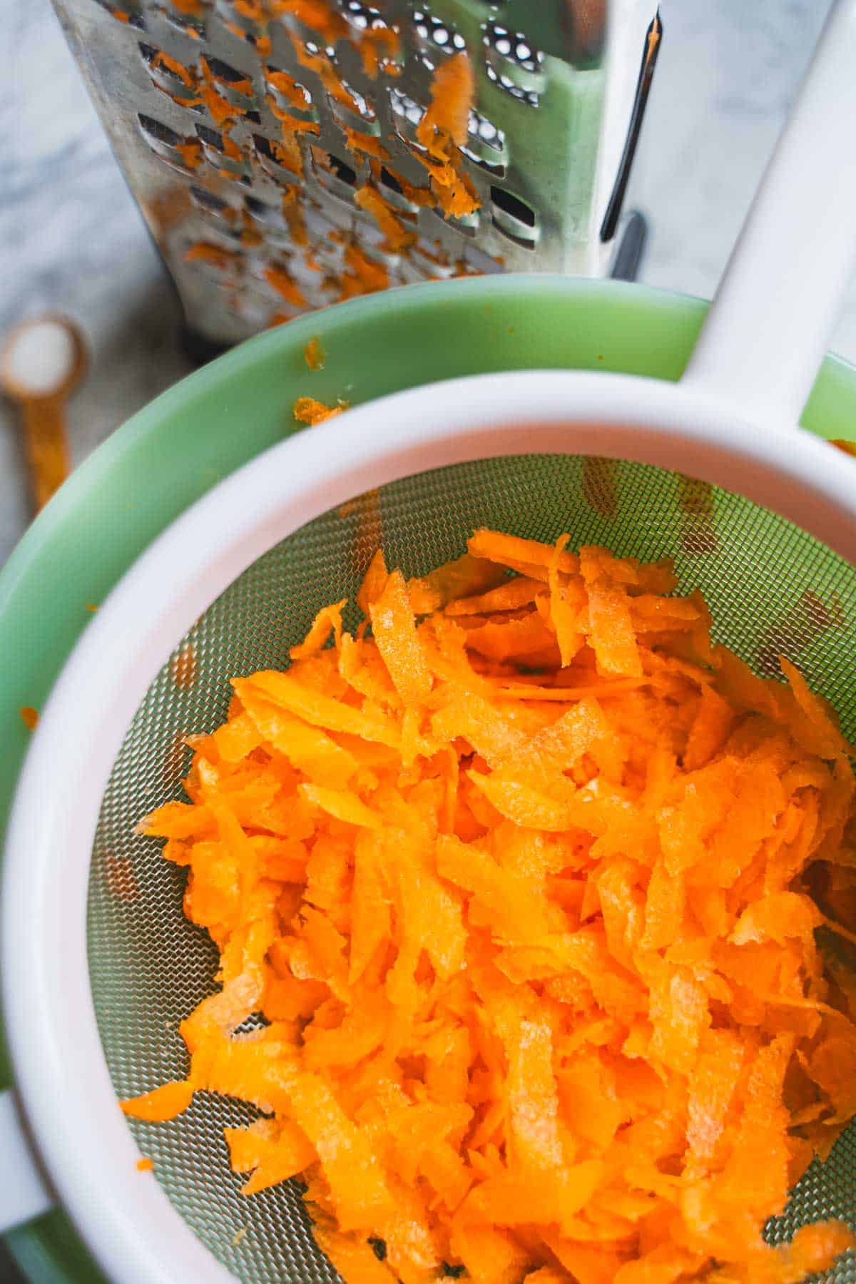Shredded carrots resting in fine mesh sieve set over a green bowl sitting next to box grater to make carrot cake cookies.