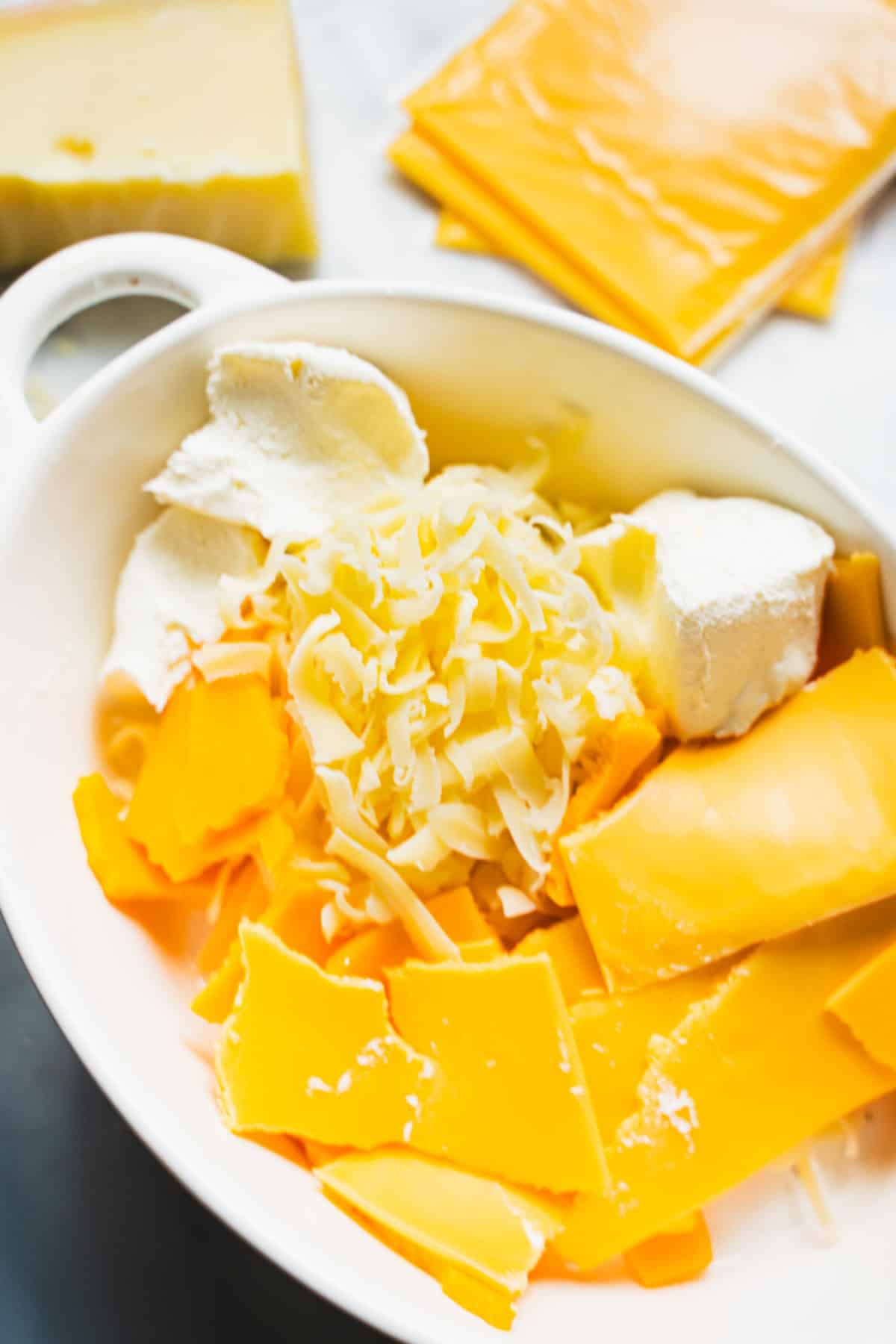 Cream cheese cubes, shredded emmental cheese, crumbled cheddar and American cheese slices in a white ramakin.