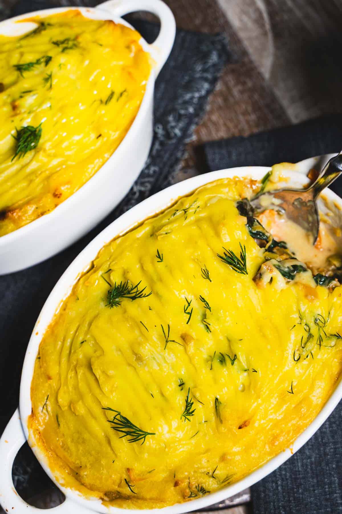 Two white ramekins of seafood pot pie or fisherman's pie on a wooden table with black fringed napkins.