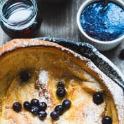 Dutch Baby Pancake Recipe for Two in a cast iron skillet garnished with blueberries and powdered sugar with blueberry sauce in white ramekin and maple syrup in a jar.