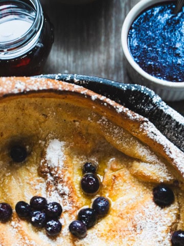 Dutch Baby Pancake Recipe for Two in a cast iron skillet garnished with blueberries and powdered sugar with blueberry sauce in white ramekin and maple syrup in a jar.